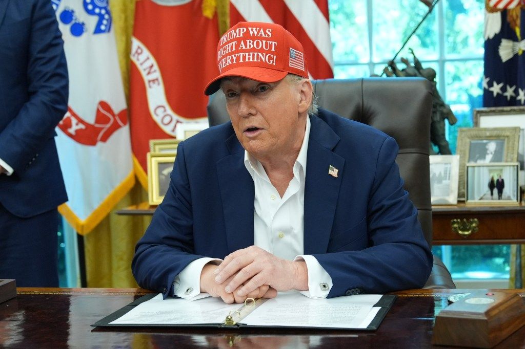 Donald Trump in the Oval Office wearing a hat that reads 
