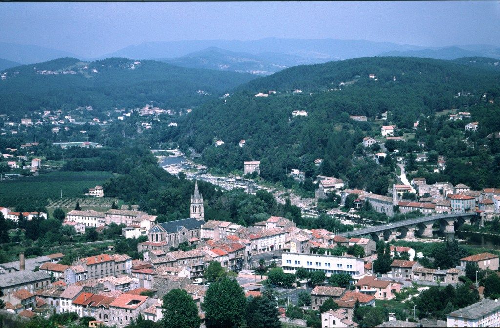 Panoramic view of a town in the Cevennes region of France, nestled in a forested river valley with a bridge connecting parts of the town.