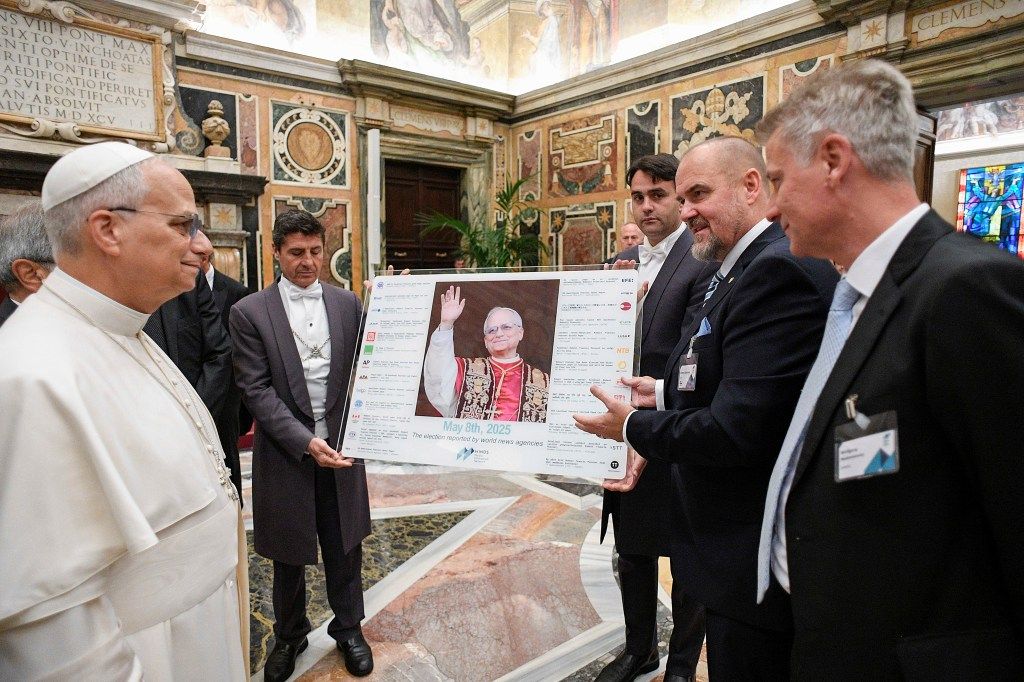 Pope Leo XIV and two men hold a placard featuring an image of Pope Leo XIV, surrounded by text about 