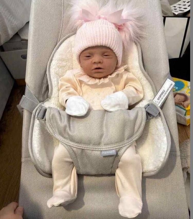 A baby doll wearing a pink hat and cream-colored clothing, lying in a baby bouncer.