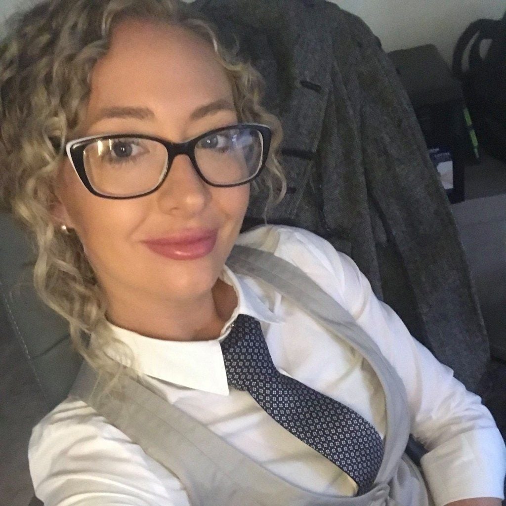 A woman with curly blonde hair and glasses wearing a white shirt, a gray vest, and a patterned
</p>
<p><a class=