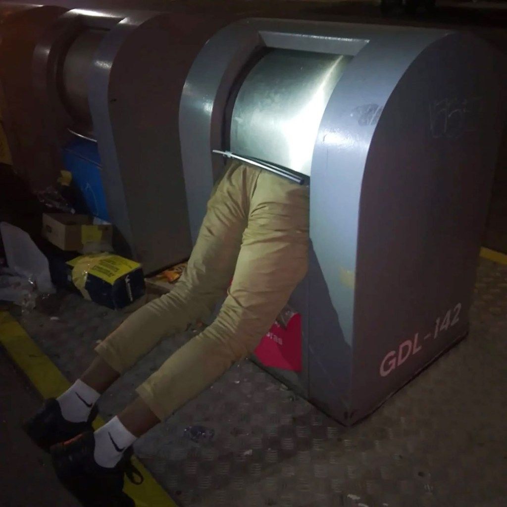 A man's lower body, dressed in khaki pants and black shoes, is seen sticking out of a grey garbage container, indicating he is stuck inside.