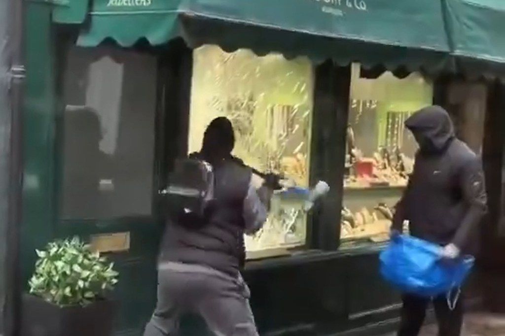 Two masked thieves robbing a jewelry store, one breaking the window with a tool, the other holding a blue bag.