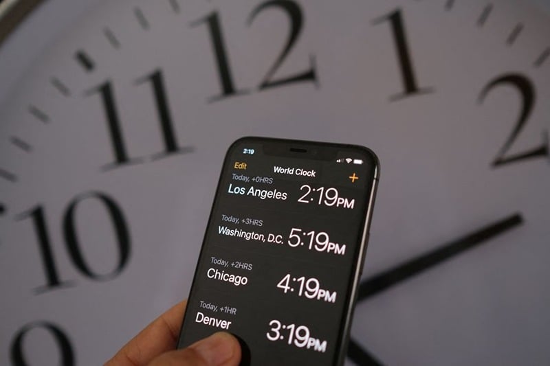 This illustration photo shows a clock in the background of a smartphone showing the time after daylight saving time was implemented in Los Angeles, California, on March 15, 2022. The US Senate advanced a bill on March 15 that would bring an end to the twice-yearly changing of clocks, in favor of a "new, permanent standard time" that would mean brighter winter evenings. (Photo by Chris DELMAS / AFP) (Photo by CHRIS DELMAS/AFP via Getty Images)