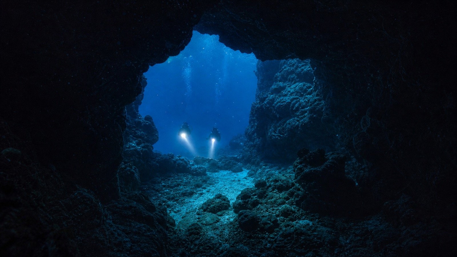 Editorial illustration of scuba divers approaching a dark underwater cave in the Maldives